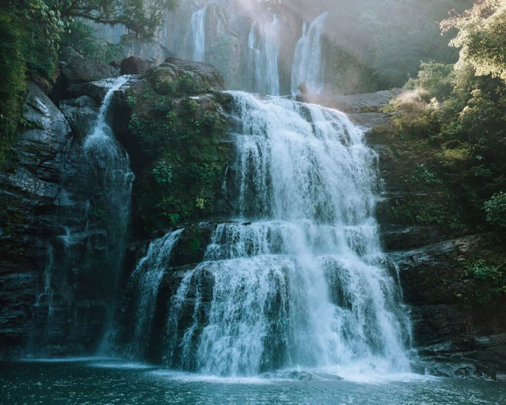 A picturesque waterfall cascading over multiple rocky levels surrounded by lush greenery, with sunlight filtering through the trees. The water flows into a serene pool, creating a tranquil nature scene.