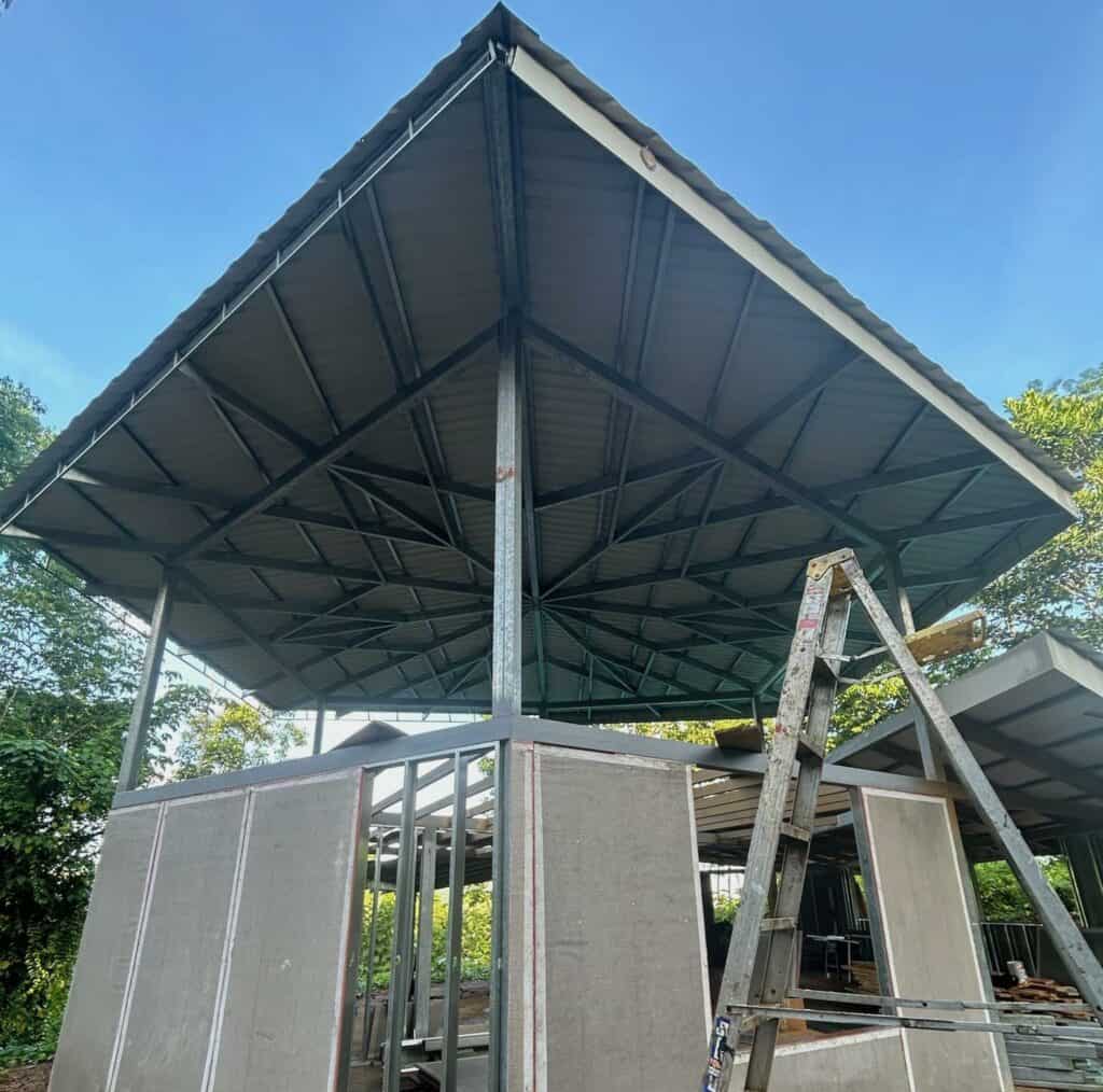 A metal structure with a large roof is under construction. A ladder leans against it. The frame is set against a backdrop of trees and blue sky.