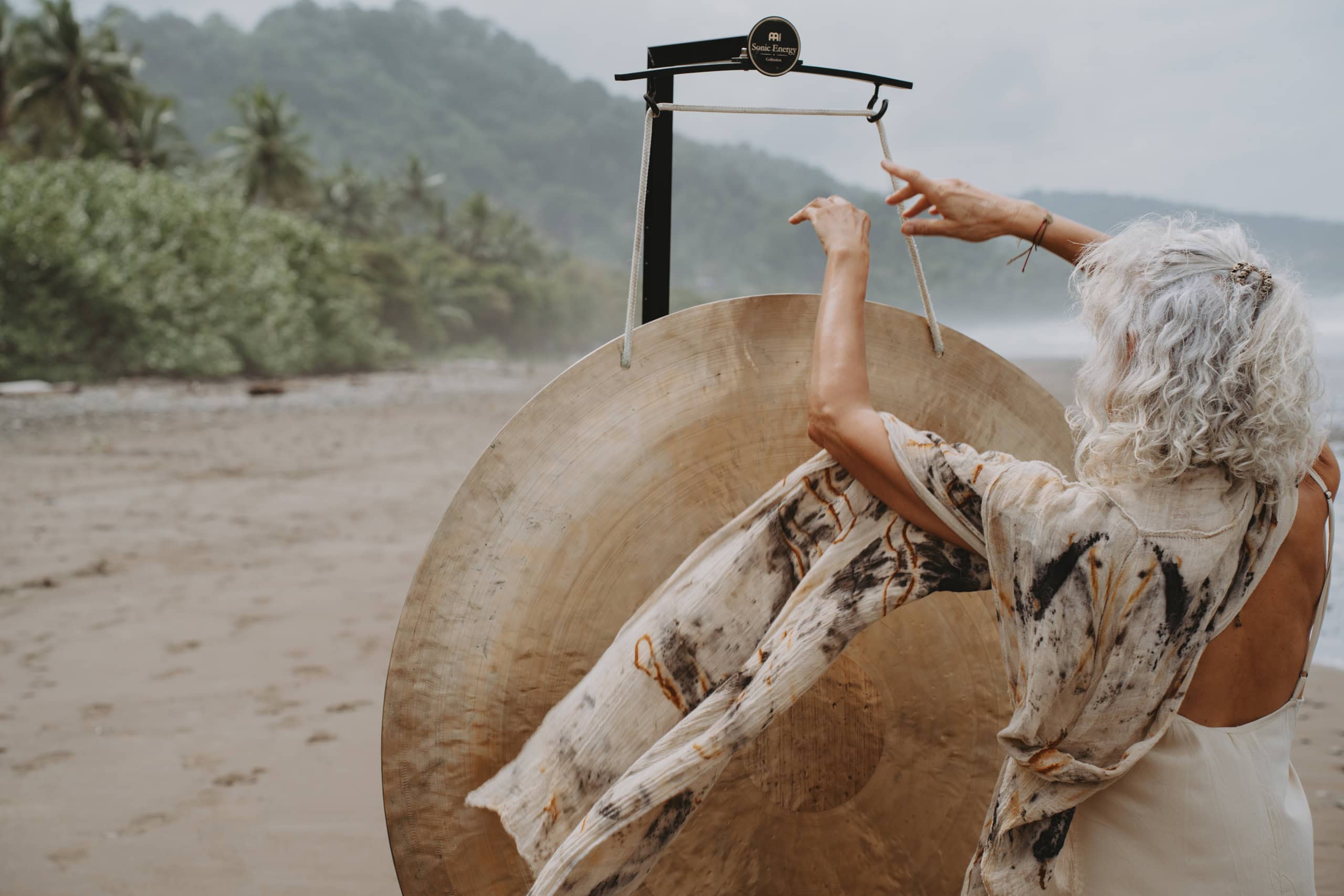 An elderly person with white hair plays a large gong on a foggy beach surrounded by lush greenery and palm trees. They wear a patterned shawl, and the atmosphere is calm and serene.