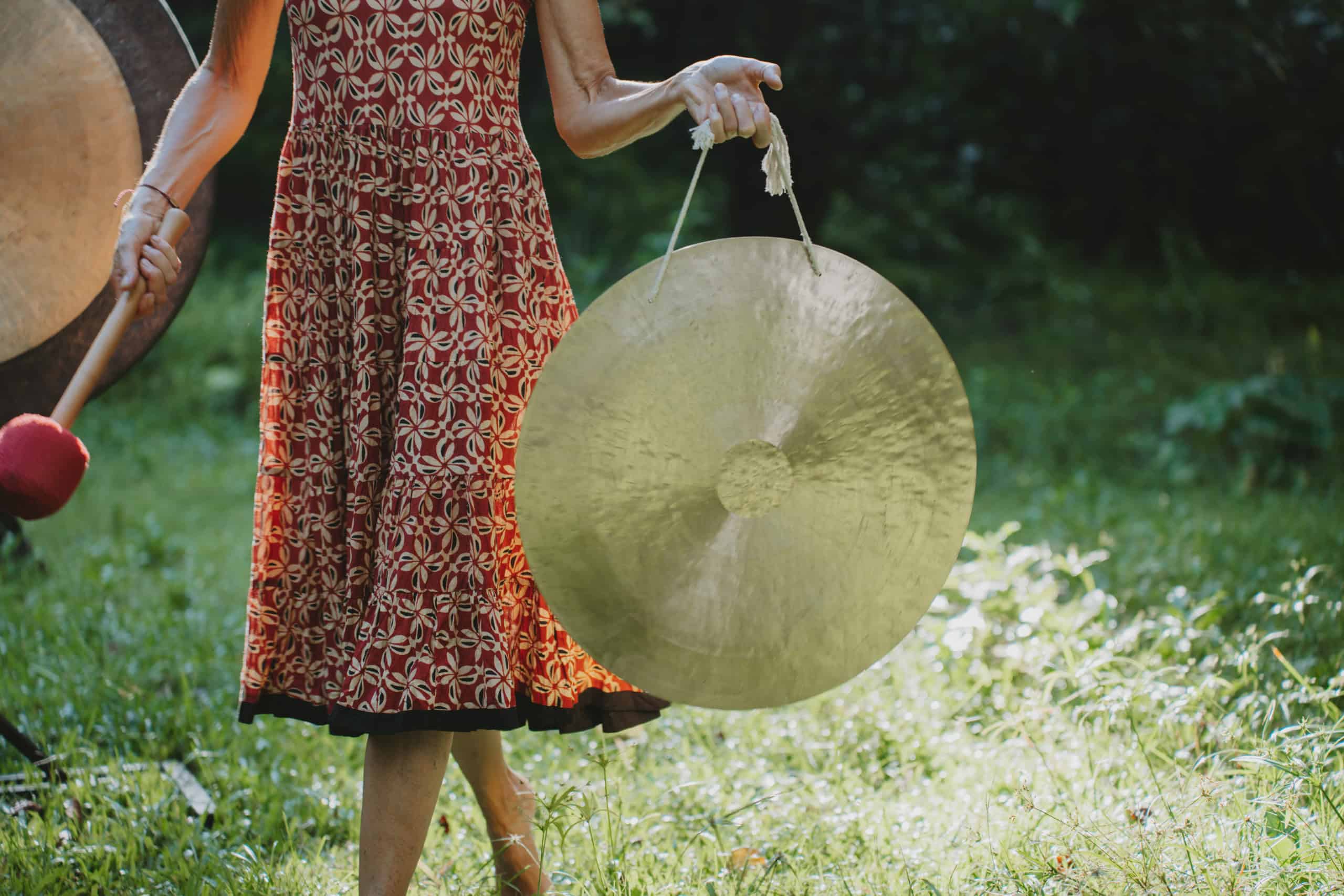 A person in a red patterned dress holds a large circular golden gong in one hand and a mallet in the other, standing on a grassy area with sunlight filtering through the trees.