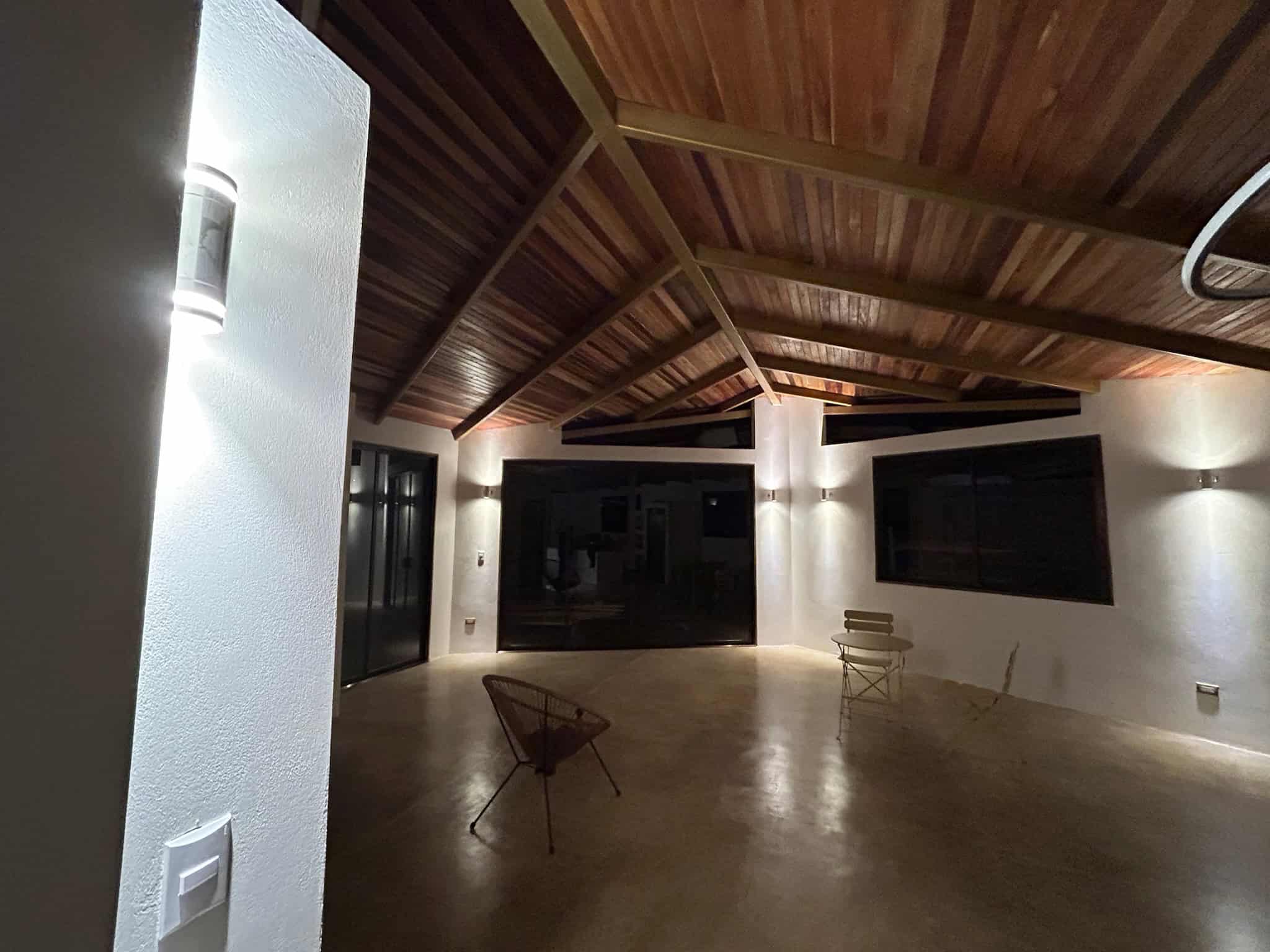 Spacious modern room with a high, wooden-beamed ceiling. The space features minimalistic decor, including two chairs and polished concrete flooring. Warm lighting highlights the contemporary style and clean lines of the architecture.