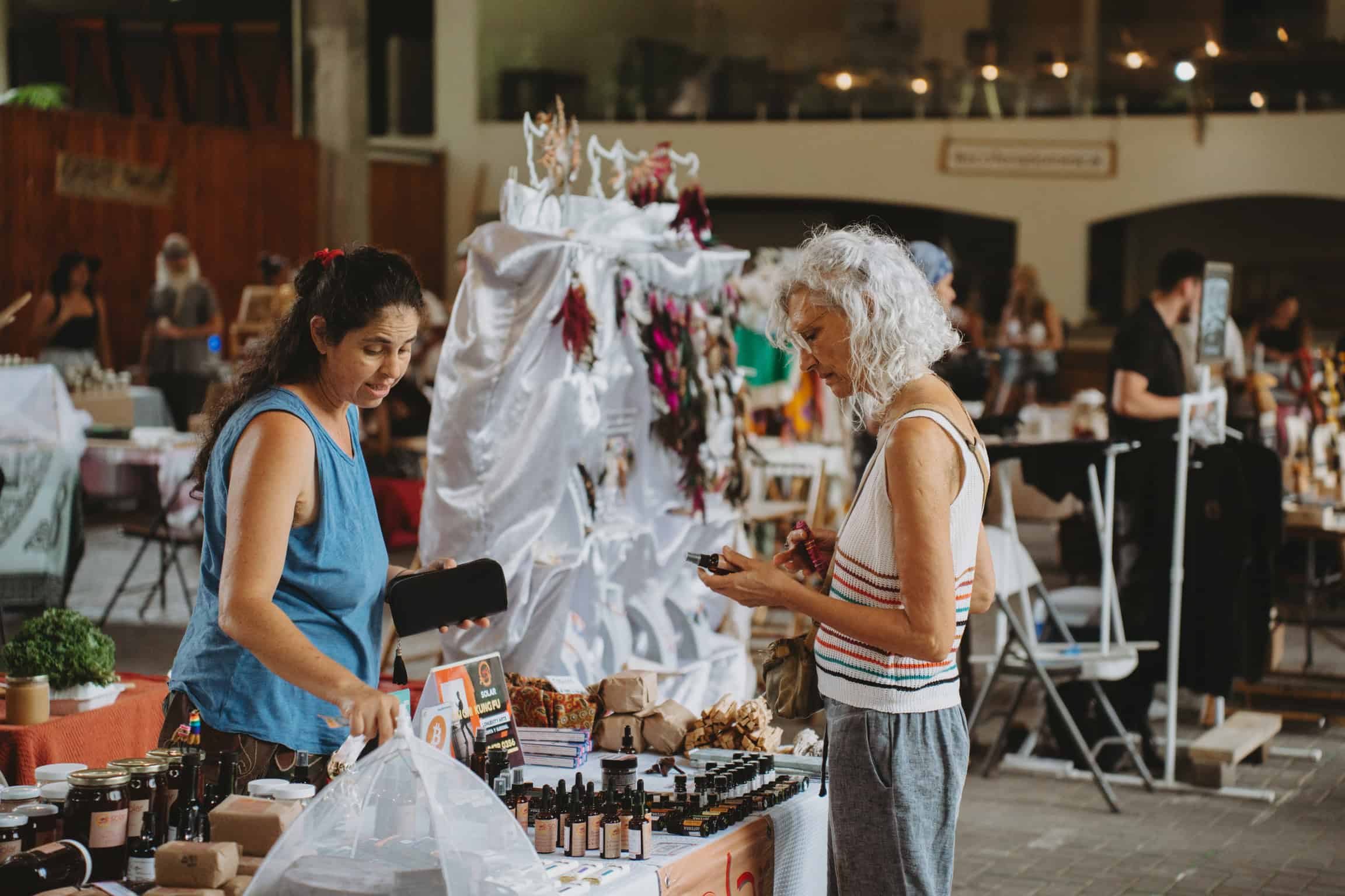 Two women are engaged at a table in a busy indoor market. One is holding a phone, while the other appears to be organizing items. The table is filled with various products and bottles. People and displays are visible in the background.